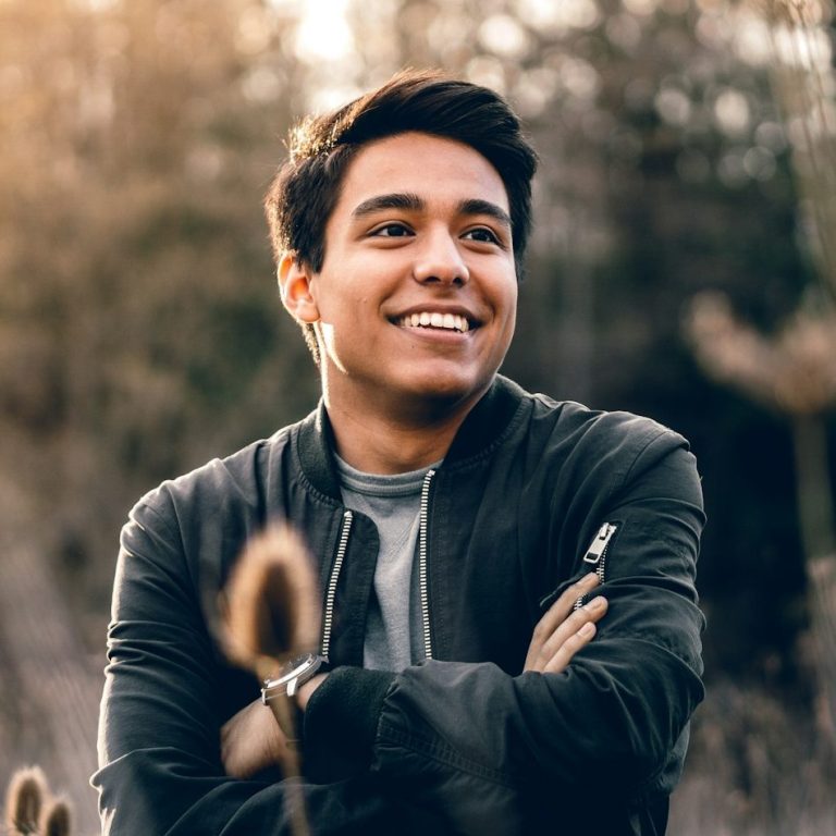 A young man smiles with arms crossed, standing in a natural setting.